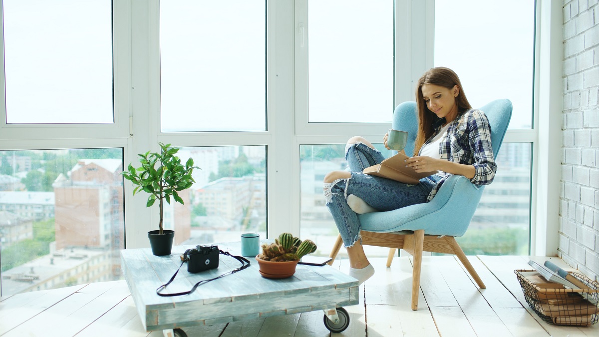 Young woman reading book and drinking coffee sitting on balcony in modern loft apartment.