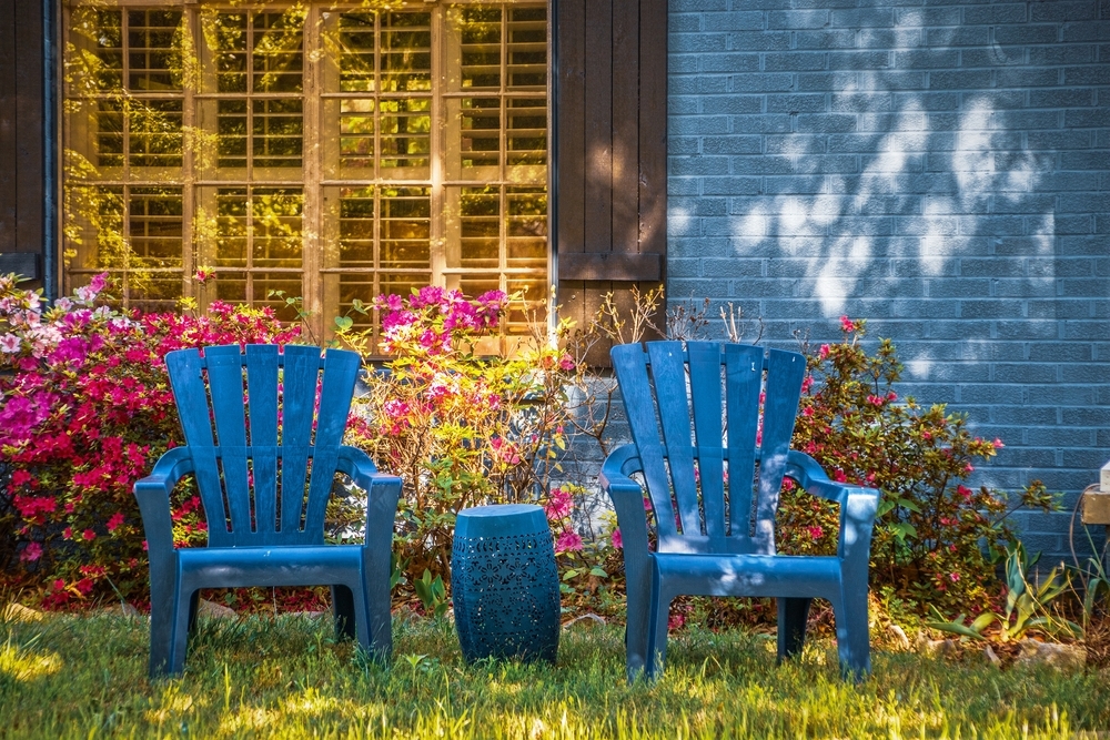 Two blue Adirondack chairs and matching drum table sitting on lawn in front of shuttered window in blue brick house landscaped with azaleas in dappled light