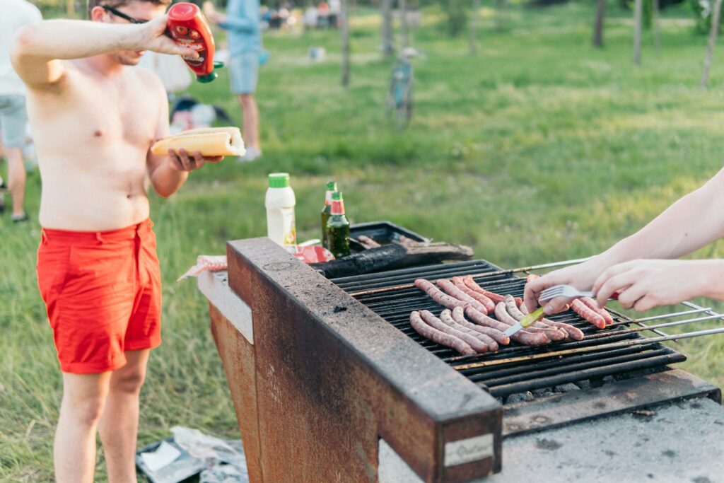 Guy putting ketchup to his hotdog next to the barbacue where there are other sausages.